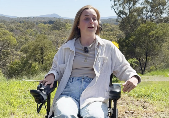 Woman in a wheelchair outdoors on a sunny day, wearing a beige top and light jacket with a sunflower nearby.