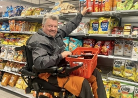 Wheelchair user in supermarket with elevated seat to reach higher shelf.